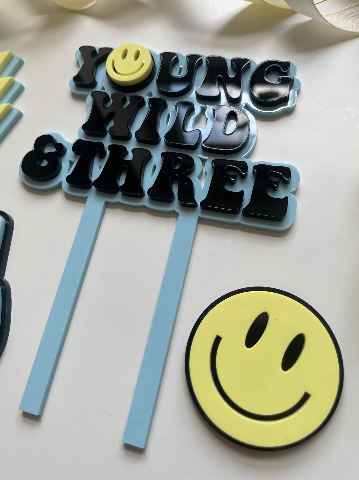 young wild and three acrylic cake topper set in blue yellow and black with lightning peace hand and smiley face charm