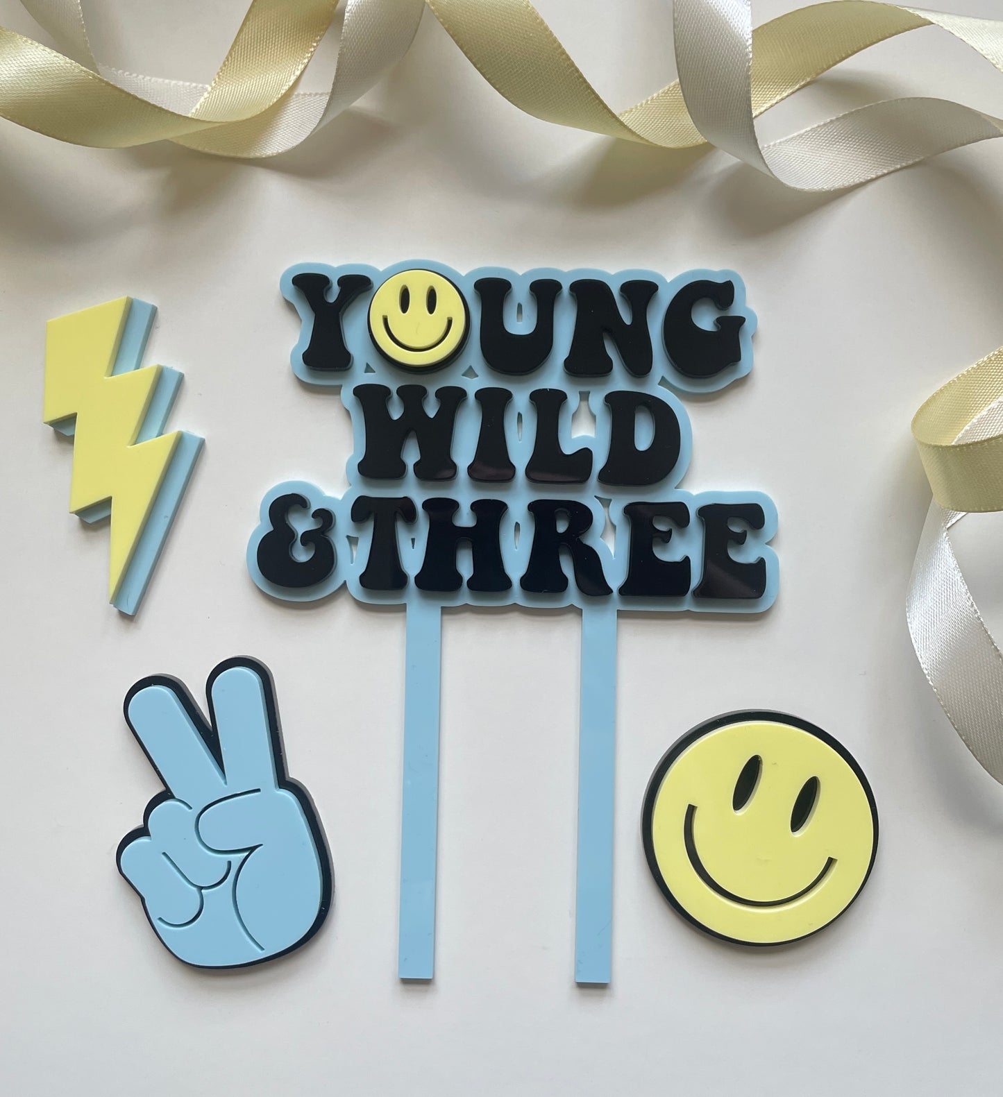 young wild and three acrylic cake topper set in blue yellow and black with lightning peace hand and smiley face charm
