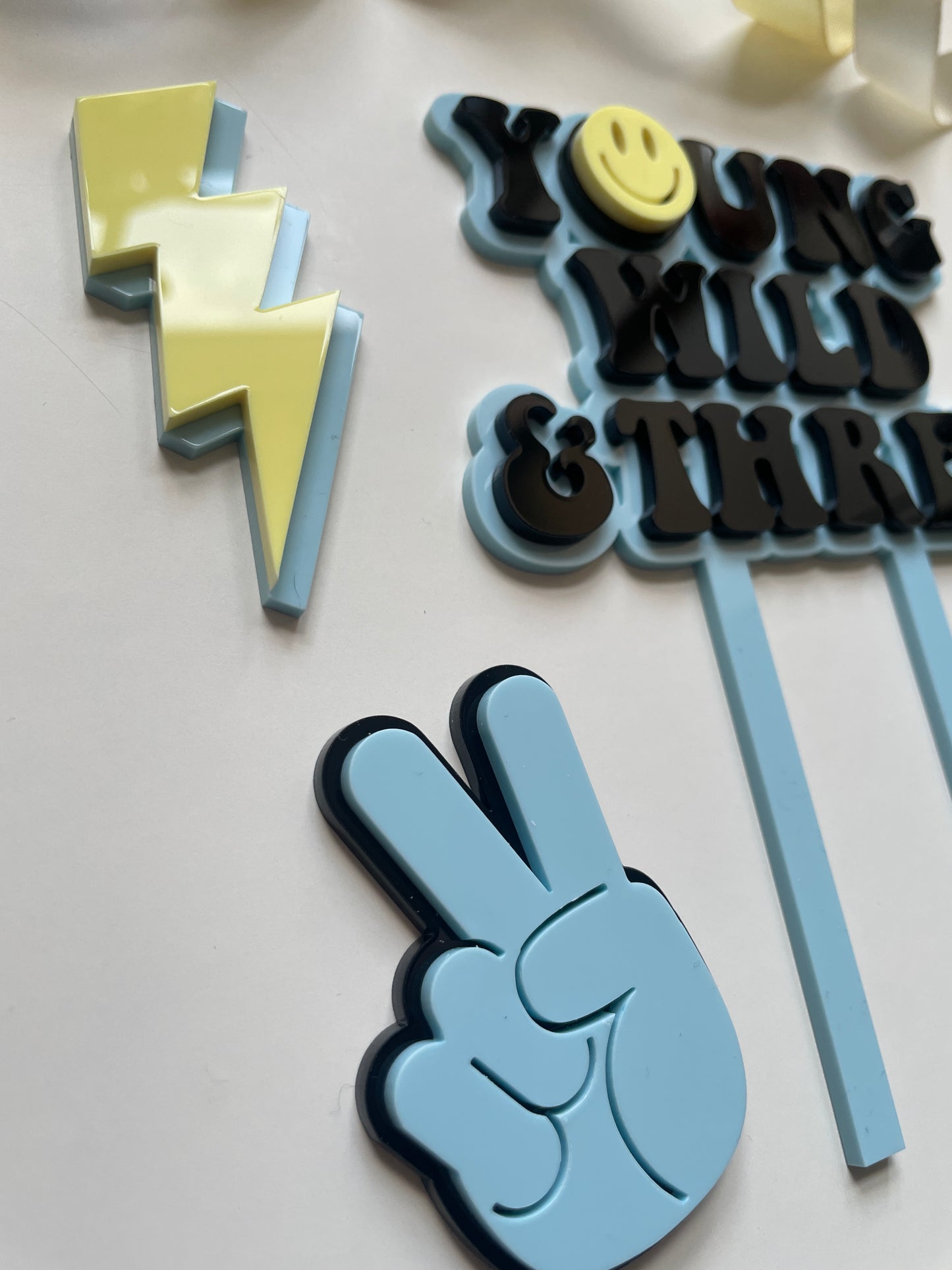 young wild and three acrylic cake topper set in blue yellow and black with lightning peace hand and smiley face charm