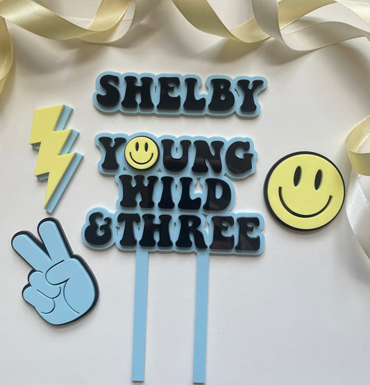 young wild and three acrylic cake topper set in blue yellow and black with lightning peace hand and smiley face charm